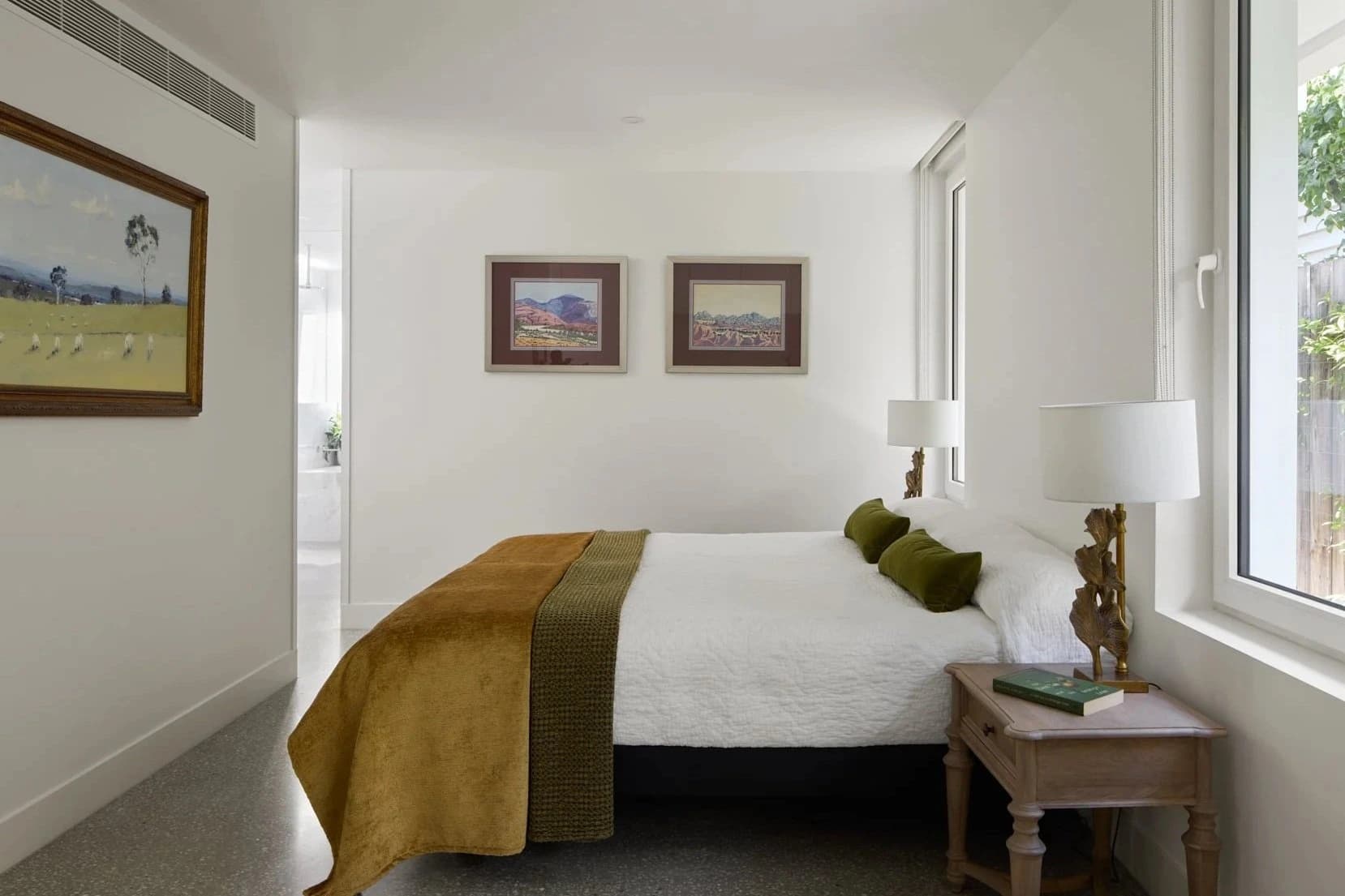 A cozy bedroom with a white bed, green pillows, wooden nightstand, white lamps, large windows, and framed landscape artwork on the white walls.