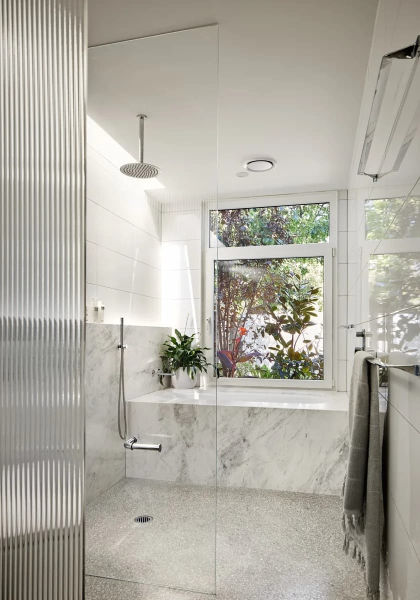 A step-free ensuite with a glass-screened shower, marble bath hob and a window onto garden foliage.