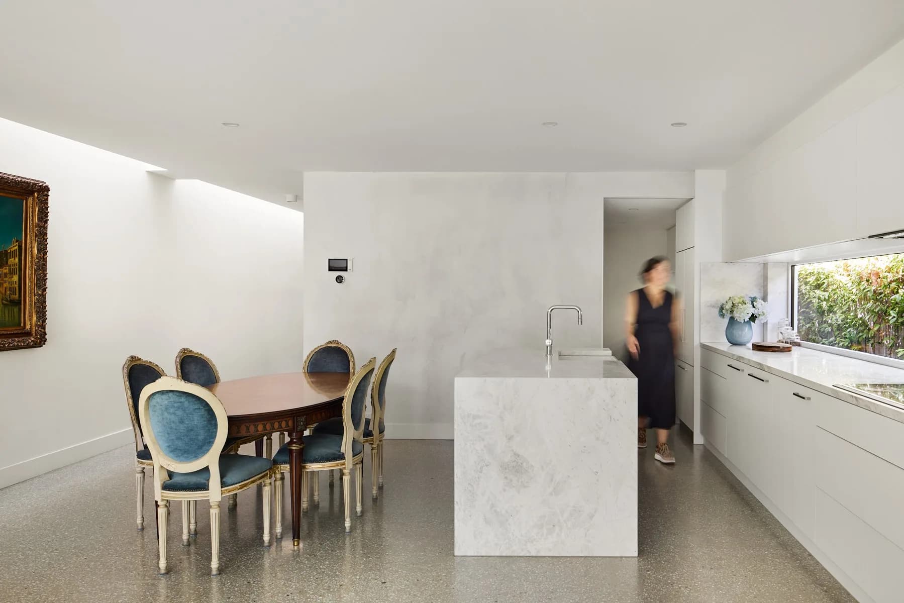 An open dining and kitchen area with an oval timber table and blue velvet chairs beside a marble island and white cabinetry.