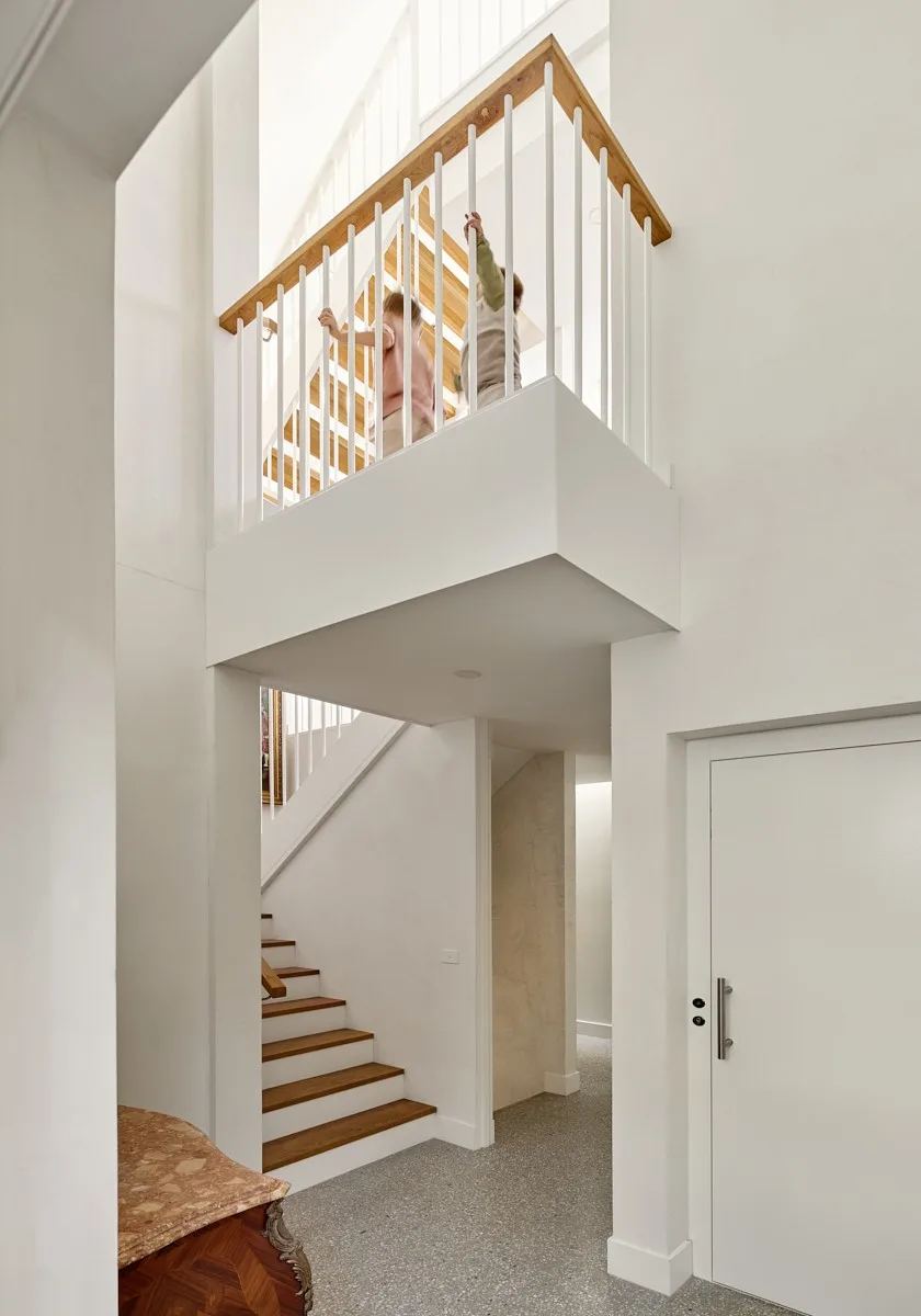 A double-height entry with an oak and white staircase, a first-floor landing where two children lean on the balustrade, and a speckled terrazzo floor.