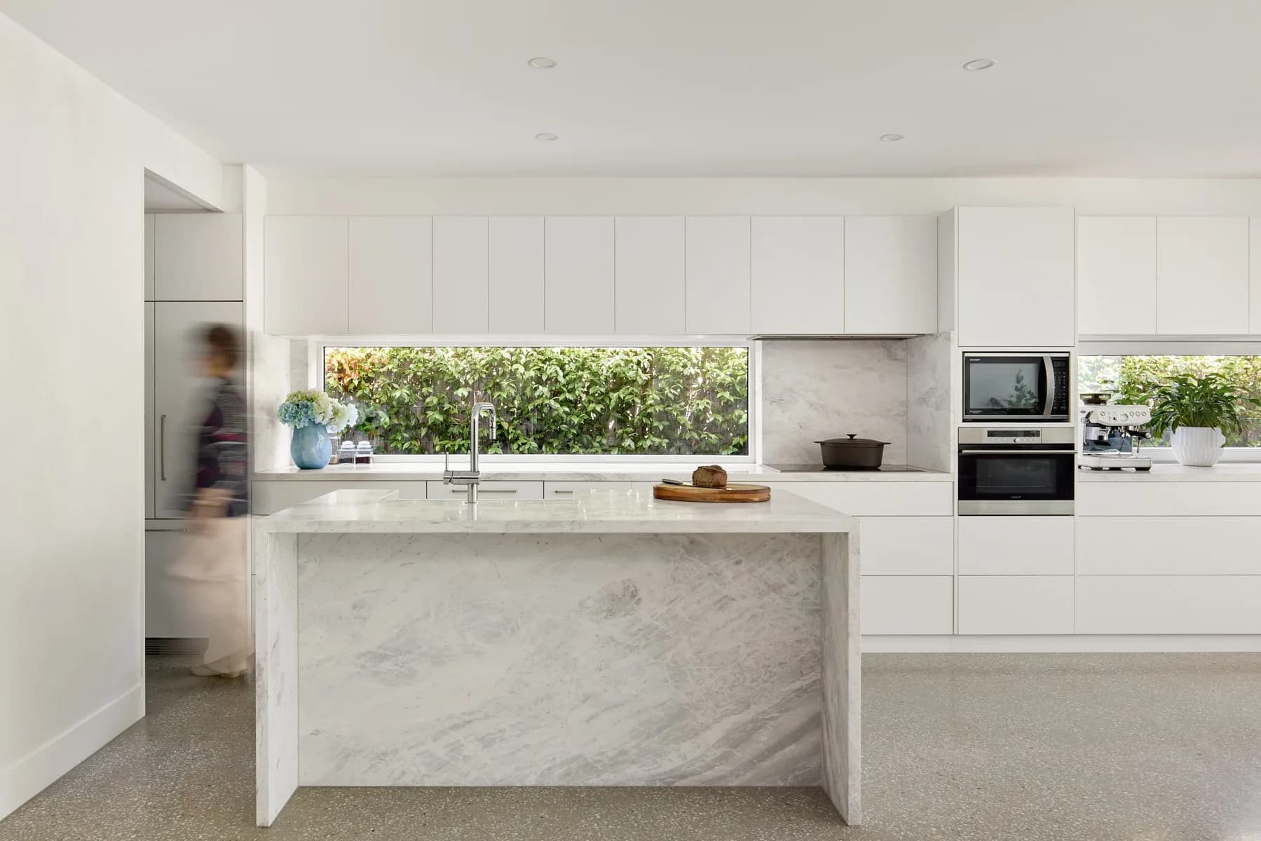 A bright white kitchen with a marble waterfall island, a long window over the bench looking onto greenery, and a blurred figure walking past.